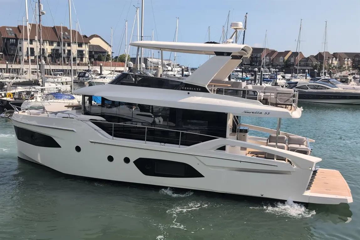 A sleek white motor yacht, labelled ‘Absolute Navetta 53’, in a marina surrounded by sailboats and moored yachts.